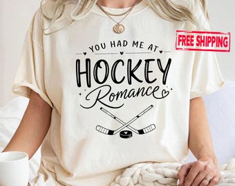 May include: A cream-coloured short-sleeve t-shirt featuring black text that reads "YOU HAD ME AT HOCKEY Romance" with small heart accents. Below the text, there is a graphic of two crossed hockey sticks and a hockey puck with a heart cutout.