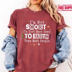 May include: A maroon short-sleeved t-shirt featuring the white text I'm Not SHORT I'm Just More Down TO EARTH Than Most People, accented with small orange stars, green and purple lightning bolts, and white sparkles on the front chest.