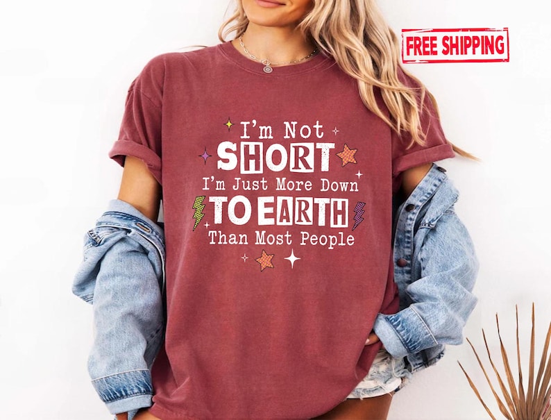 May include: A maroon short-sleeved t-shirt featuring the white text I'm Not SHORT I'm Just More Down TO EARTH Than Most People, accented with small orange stars, green and purple lightning bolts, and white sparkles on the front chest.