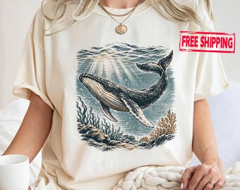 May include: A cream-colored short-sleeve t-shirt featuring a detailed graphic illustration of a humpback whale swimming underwater. The design includes light rays filtering through the water, coral, and rocky textures in shades of blue and brown.