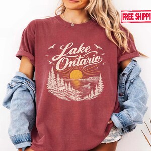 May include: A maroon short-sleeve t-shirt featuring a graphic print of Lake Ontario with a yellow sun, pine trees, a moose, and birds. The text Lake Ontario is printed in a white script font above the sun and landscape illustration.