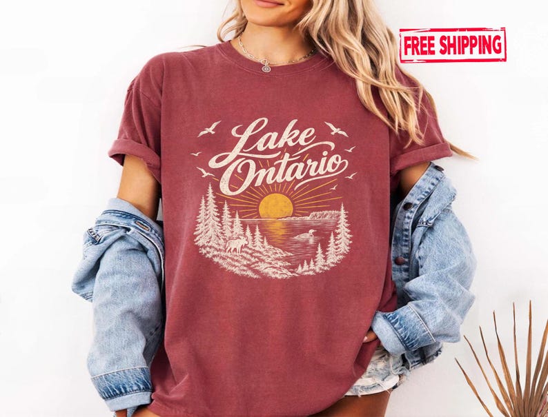 May include: A maroon short-sleeve t-shirt featuring a graphic print of Lake Ontario with a yellow sun, pine trees, a moose, and birds. The text Lake Ontario is printed in a white script font above the sun and landscape illustration.