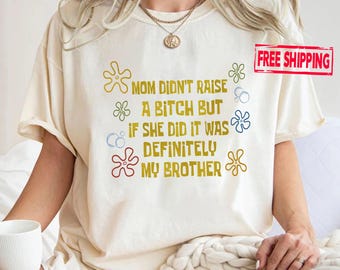 May include: A cream-colored short-sleeve t-shirt featuring the text "MOM DIDN'T RAISE A BITCH BUT IF SHE DID IT WAS DEFINITELY MY BROTHER" in mustard yellow, surrounded by small, colorful flower and bubble graphics in red, blue, and green.