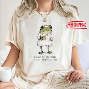 May include: A cream-colored t-shirt featuring a graphic of a green frog dressed in a white nurse uniform and cap, holding a small mug. Below the frog, the text reads: I Have to ask weird stuff or you'll die. The shirt has short sleeves and a crew neck.