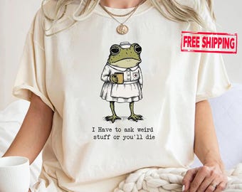 May include: A cream-coloured t-shirt featuring a graphic of a green frog dressed in a white nurse uniform and cap, holding a small mug. Below the frog, the text reads: I Have to ask weird stuff or you'll die. The shirt has short sleeves and a crew neck.
