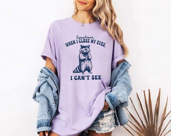 Könnte beinhalten: Ein hellviolettes T-Shirt mit kurzen Ärmeln und einer dunkelblauen Grafik eines Waschbären, der mit verschränkten Armen steht. Der Text über dem Waschbären lautet "Sometimes, WHEN I CLOSE MY EYES," und darunter "I CAN'T SEE."