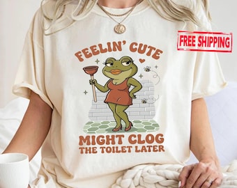 May include: A cream-colored t-shirt featuring a graphic of a green frog wearing a brown dress and holding a plunger. The text reads "FEELIN' CUTE MIGHT CLOG THE TOILET LATER" in brown letters. The design includes small bees and a brick wall background.