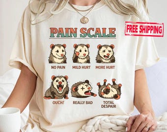 May include: Cream-colored t-shirt featuring a humorous "Pain Scale" graphic with opossums. The scale ranges from "No Pain" to "Total Despair." The text includes "Ouch!" and "Really Bad."
