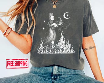 May include: A charcoal grey short-sleeve t-shirt featuring a white graphic print of a figure in a pointed hat tied to a post surrounded by flames, with a crescent moon and stars in the background. The design is centred on the chest of the shirt.