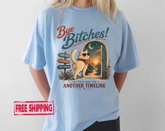 Könnte beinhalten: Ein hellblaues T-Shirt mit einer Grafik einer Katze mit Sonnenbrille und rotem Halstuch vor einer offenen Tür. Der Text lautet "Bye Bitches!" und "Switching to another timeline," mit Wegweisern "New Vibes," "Better Days," und "New Timeline."