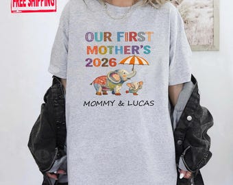 May include: A light gray t-shirt with the text "OUR FIRST MOTHER'S 2026" in colorful letters. Below the text is an illustration of two elephants under an umbrella, with the words "MOMMY & LUCAS" underneath.
