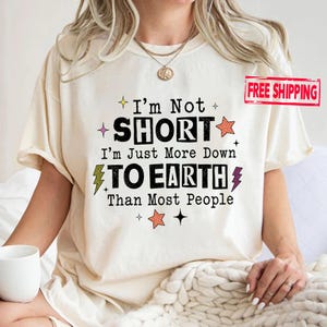 May include: A cream-colored short-sleeve t-shirt featuring black text that reads I'm Not SHORT I'm Just More Down TO EARTH Than Most People. The design includes small orange and purple lightning bolts and star icons surrounding the printed text.