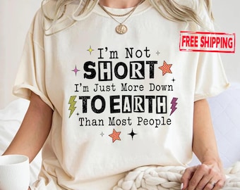 May include: A cream-colored short-sleeve t-shirt featuring black text that reads I'm Not SHORT I'm Just More Down TO EARTH Than Most People. The design includes small orange and purple lightning bolts and star icons surrounding the printed text.