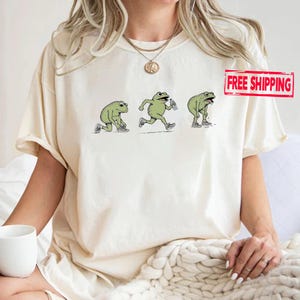 May include: A cream-colored t-shirt featuring a graphic illustration of a green frog in three stages of exercise: tying a sneaker, running while holding a water bottle, and standing while panting. The frog wears white sneakers in each pose.