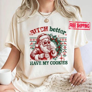 May include: A cream-colored t-shirt featuring a graphic of Santa Claus wearing sunglasses, holding a cookie and a mug. The text reads "BITCH better HAVE MY COOKIES" in red and green. The design includes a festive pattern with holly and reindeer.