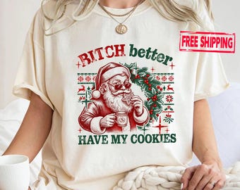May include: A cream-coloured t-shirt featuring a graphic of Santa Claus wearing sunglasses, holding a cookie and a mug. The text reads "BITCH better HAVE MY COOKIES" in red and green. The design includes a festive pattern with holly and reindeer.