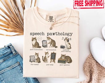 May include: Cream-colored t-shirt with a graphic featuring cartoon cats illustrating speech therapy terms like "purr-nunciation" and "cat-iculation." The shirt has a crew neck and a small tag. The words "speech pawthology" are printed at the top.
