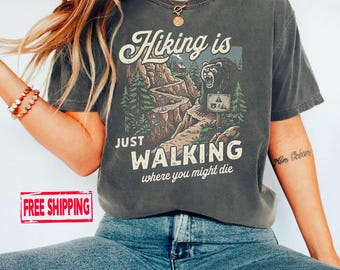 May include: Dark grey t-shirt with the text "Hiking is Just Walking Where You Might Die" and a graphic of a bear and a hiking trail. The design includes trees and a sign. The shirt has a relaxed fit.