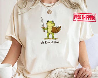 May include: Cream-colored t-shirt featuring a cartoon frog holding a sword and a book. The frog is green with a yellow belly. The text "We Read at Dawn!" is printed below the frog in black.