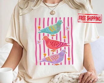 May include: A cream-colored t-shirt featuring a graphic print of three pigeons in teal, pink, and purple, layered over vertical pink stripes. The design includes small yellow stars and purple flowers scattered around the birds on the front.