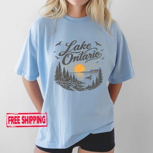 May include: A light blue short-sleeve t-shirt featuring a dark brown graphic print of a lake landscape with pine trees, a sun, birds, and the text Lake Ontario. The design includes a small animal on the shore and a bird swimming in the water.