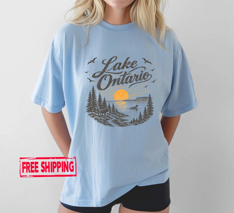 May include: A light blue short-sleeve t-shirt featuring a dark brown graphic print of a lake landscape with pine trees, a sun, birds, and the text Lake Ontario. The design includes a small animal on the shore and a bird swimming in the water.