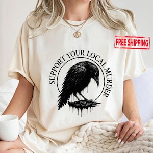 May include: Cream-colored t-shirt featuring a black raven illustration within a circular design. The text "SUPPORT YOUR LOCAL MURDER" encircles the raven. The shirt is a casual wear item, suitable for everyday use.