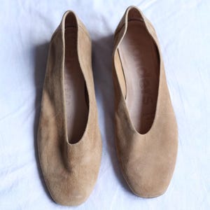 May include: A pair of tan suede flats with a rounded toe and a low-cut vamp. The shoes are displayed on a white surface. The interior of the shoe is a lighter shade of tan. The shoes appear to be unworn.