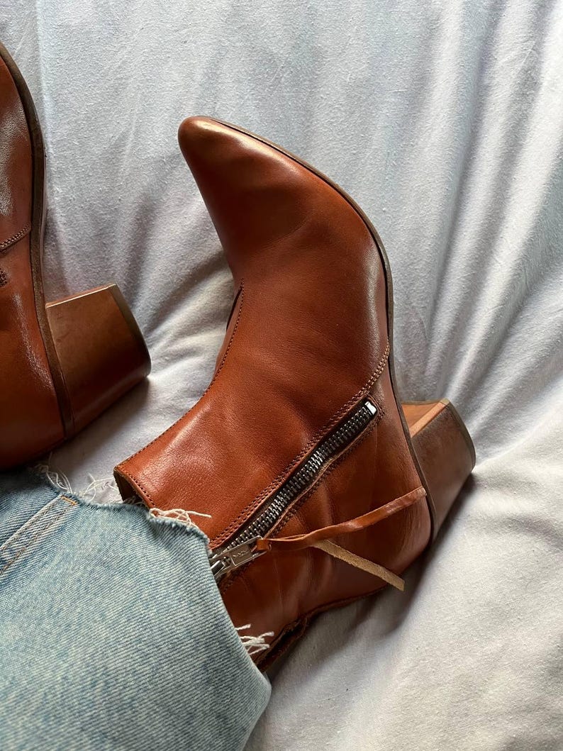 Puede incluir: Un par de botines de cuero marr&oacute;n con punta puntiaguda y cremallera en la parte posterior. Las botas tienen un tac&oacute;n de bloque y un detalle de borla.