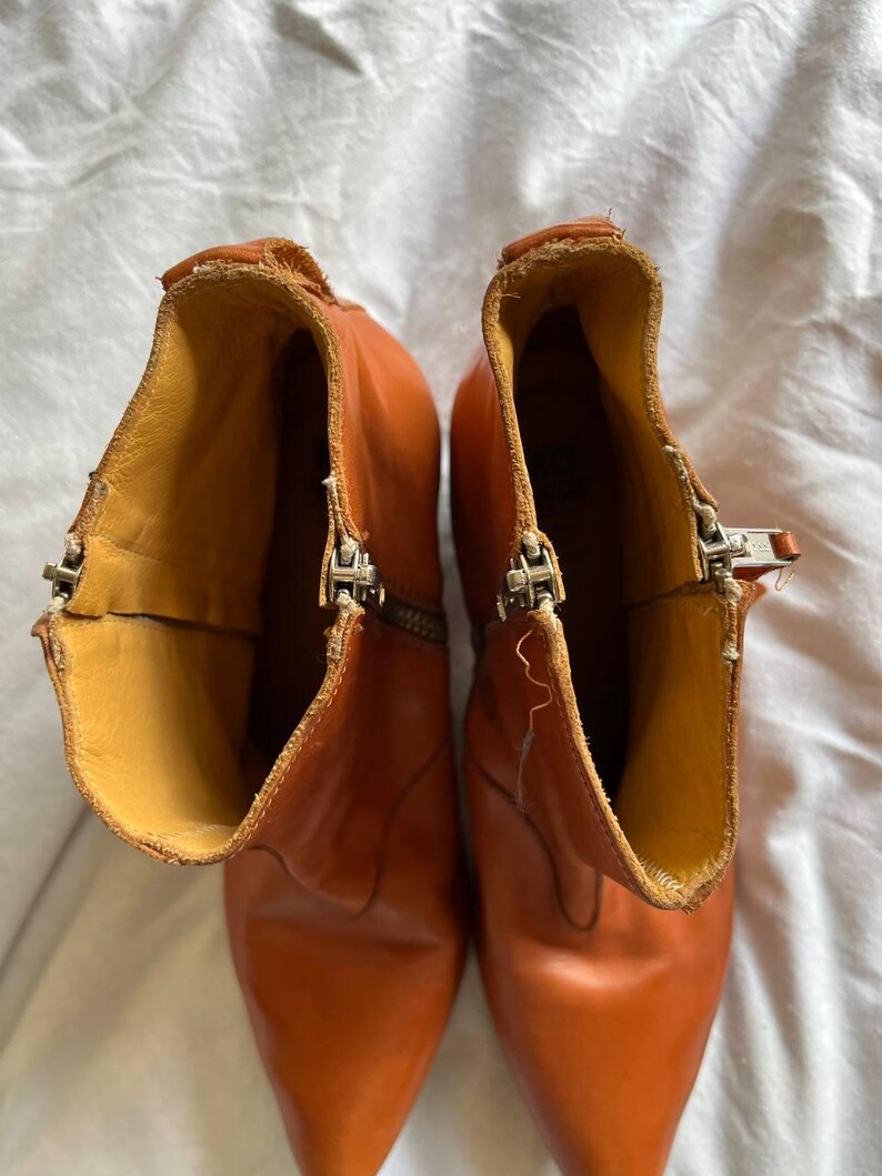 Puede incluir: Un par de botines de cuero marr&oacute;n con punta puntiaguda y cremalleras laterales. Las botas tienen un aspecto desgastado y est&aacute;n hechas de cuero suave y flexible.