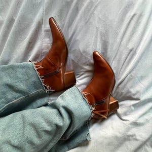Puede incluir: Un par de botas vaqueras de cuero marr&oacute;n con punta puntiaguda y tac&oacute;n de bloque. Las botas se llevan con pantalones vaqueros de mezclilla azul.