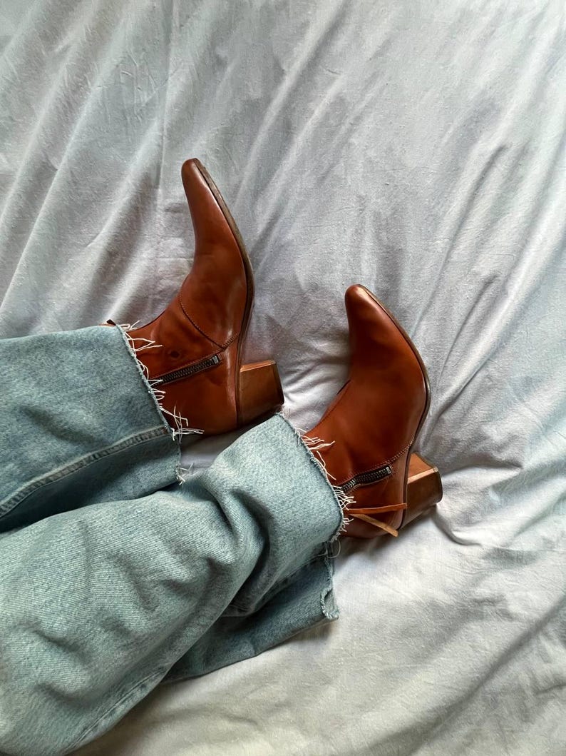 Puede incluir: Un par de botas vaqueras de cuero marr&oacute;n con punta puntiaguda y tac&oacute;n de bloque. Las botas se llevan con pantalones vaqueros de mezclilla azul.