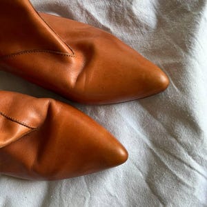Puede incluir: Un par de botines de cuero marr&oacute;n con punta puntiaguda. Los botines est&aacute;n sobre una superficie de tela blanca.