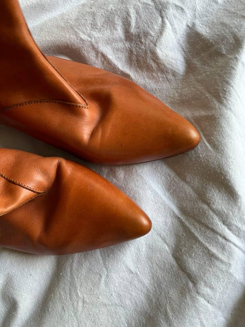 Puede incluir: Un par de botines de cuero marr&oacute;n con punta puntiaguda. Los botines est&aacute;n sobre una superficie de tela blanca.