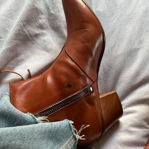 Puede incluir: Botas de tobillo de cuero marr&oacute;n con punta puntiaguda y cremallera en la parte posterior. Las botas tienen un tac&oacute;n de bloque y se llevan con jeans azules.