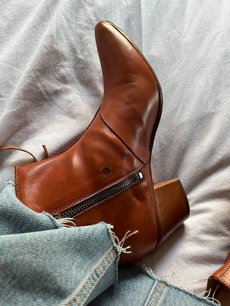 Puede incluir: Botas de tobillo de cuero marr&oacute;n con punta puntiaguda y cremallera en la parte posterior. Las botas tienen un tac&oacute;n de bloque y se llevan con jeans azules.