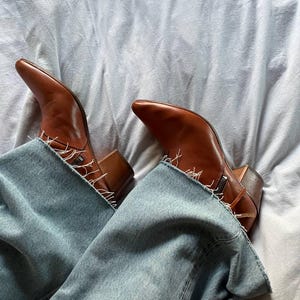 Puede incluir: Un par de botas vaqueras de cuero marr&oacute;n con punta puntiaguda y un detalle de cremallera en el lateral. Las botas se llevan con unos vaqueros azules.