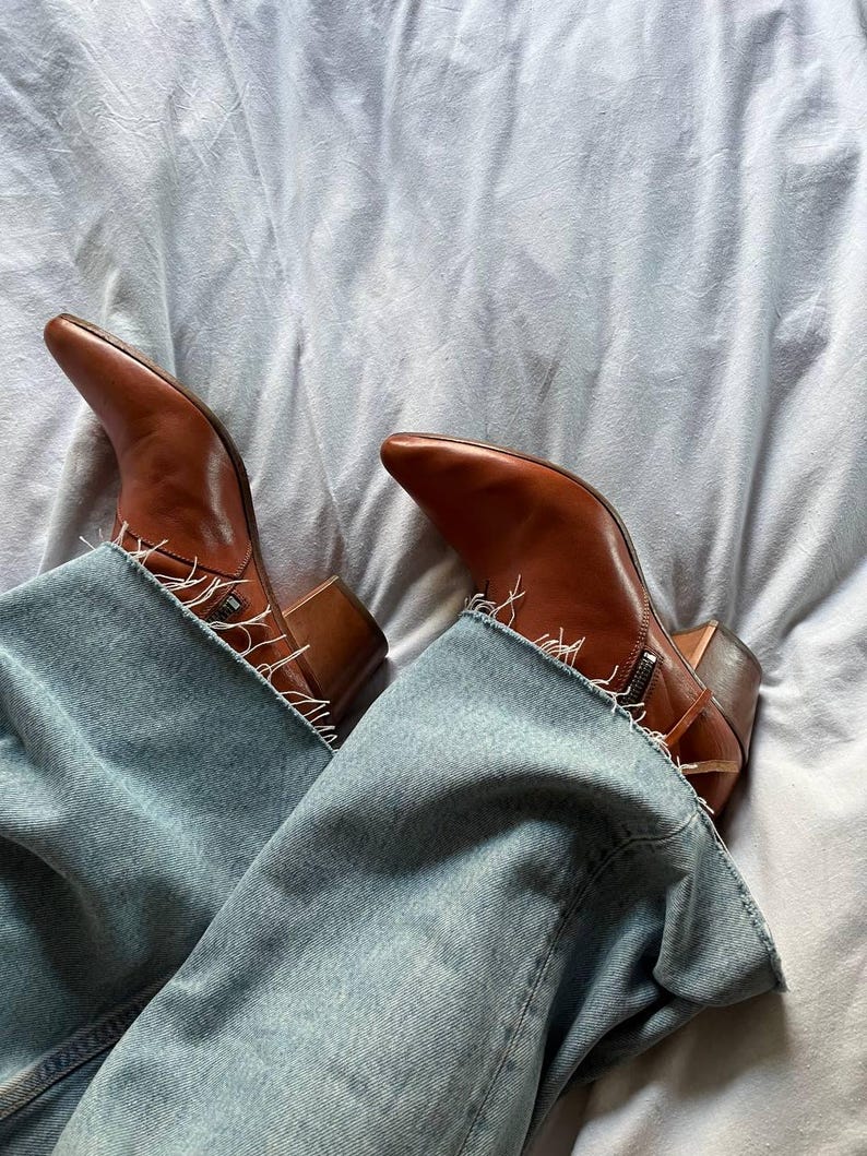 Puede incluir: Un par de botas vaqueras de cuero marr&oacute;n con punta puntiaguda y un detalle de cremallera en el lateral. Las botas se llevan con unos vaqueros azules.
