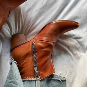 Puede incluir: Botas de tobillo de cuero marr&oacute;n con cremallera lateral. Las botas se llevan con vaqueros azules.