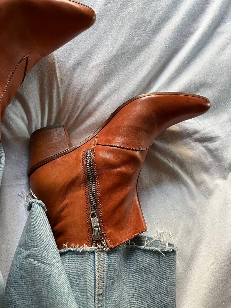Puede incluir: Botas de tobillo de cuero marr&oacute;n con cremallera lateral. Las botas se llevan con vaqueros azules.