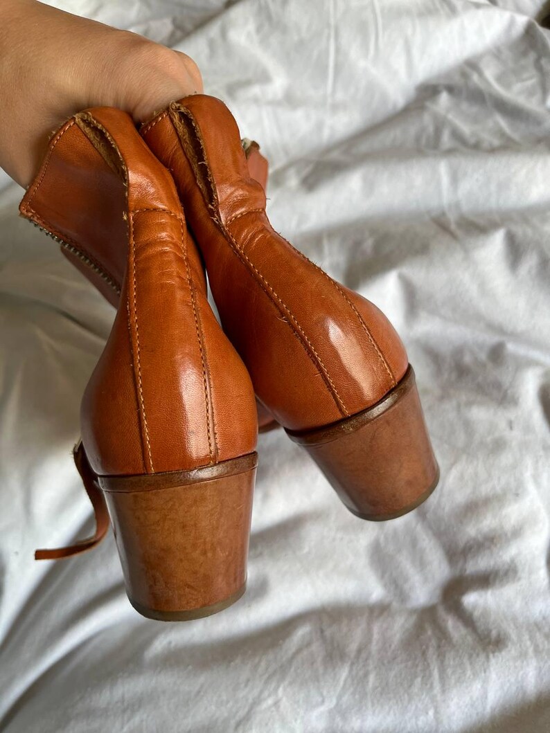 Puede incluir: Un par de botines de cuero marr&oacute;n con tac&oacute;n grueso. Las botas tienen una cremallera en el interior de cada bota y una correa alrededor del tobillo.