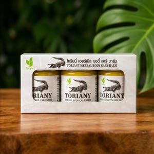 Könnte beinhalten: Ein rechteckiger weißer Karton mit drei kleinen Glastiegeln Toriany Herbal Body Care Balm. Jeder Tiegel hat ein Etikett mit einer schwarzen Krokodilillustration und dem Text Toriany Herbal Body Care Balm in schwarzer Schrift.