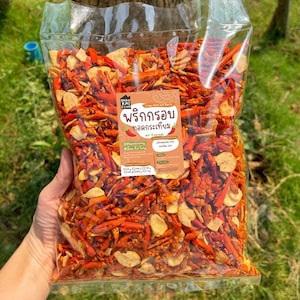 Może przedstawiać: Przezroczysta plastikowa torba wypełniona czerwonymi papryczkami chili i smażonym czosnkiem. Torba ma brązową etykietę z tekstem w obcym języku. Zawartość to mieszanka czerwieni i brązu, z jaśniejszymi chipsami czosnkowymi.