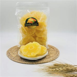 May include: A transparent bag of dried pineapple rings, labelled "Happy Fruits". Several slices are on a small white plate, placed on a woven mat. The dried fruit is a vibrant yellow colour. The image also includes some wheat.