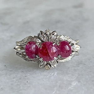 May include: A silver ring with three oval, vivid red gemstones. The ring has a floral design with small accent diamonds. The gemstones are the focal point, set against the detailed silverwork.