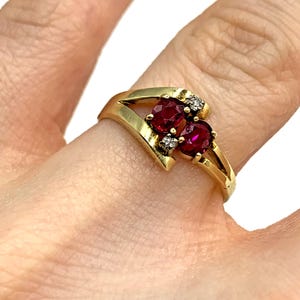 May include: A gold ring with two oval, deep red gemstones and small diamond accents. The ring has a split shank design and is set in a yellow gold band. The gemstones are the focal point, creating a classic and elegant appearance.