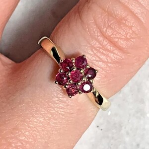 May include: A gold ring with a cluster of small, round, deep red gemstones. The gemstones are arranged in a floral pattern, with a polished gold band. The ring is shown on a finger.