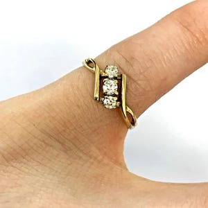 May include: A gold ring featuring three clear gemstones. The ring has a distinctive design with a curved band and a rectangular setting for the gemstones. The ring is displayed on a finger.