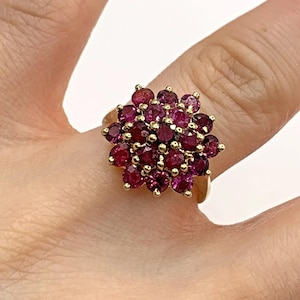 May include: A gold ring with a cluster of round, deep red gemstones. The gemstones are arranged in a floral pattern, creating a sparkling design. The ring's band is a simple, polished gold.
