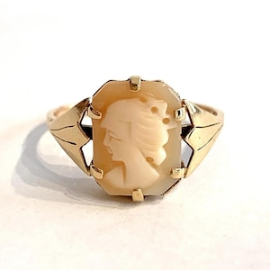 May include: A gold ring featuring a rectangular, light-coloured cameo carved with a profile of a person's head. The cameo is set in a gold frame with four prongs, and the band has decorative, angular gold shoulders on each side of the setting.